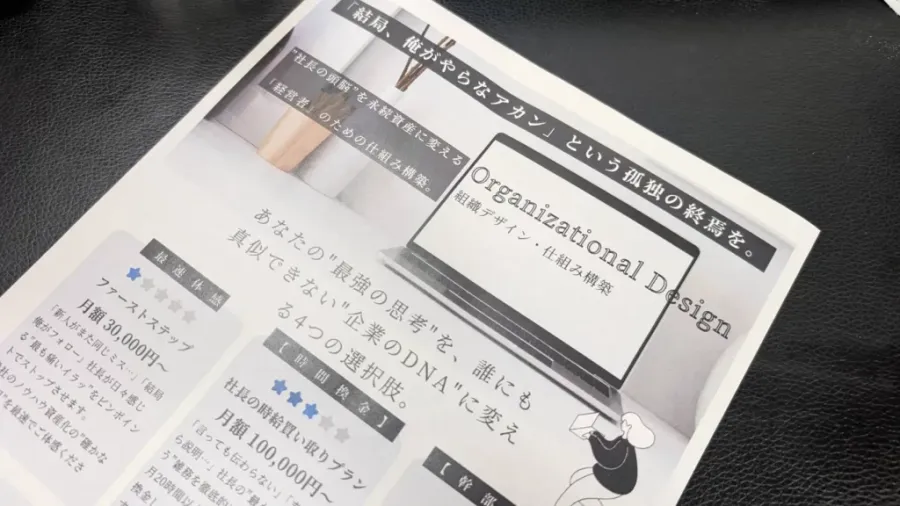 机の上に置かれた紙のチラシ。大きな見出しに「『結局、俺がやらなアカン』という孤独の終焉。」、その下に「“社長の頭脳”を永続資産に変える」「“経営者”のための仕組み構築」「Organizational Design 組織デザイン・仕組み構築」と書かれている。左側に月額のプランが並んでいる。